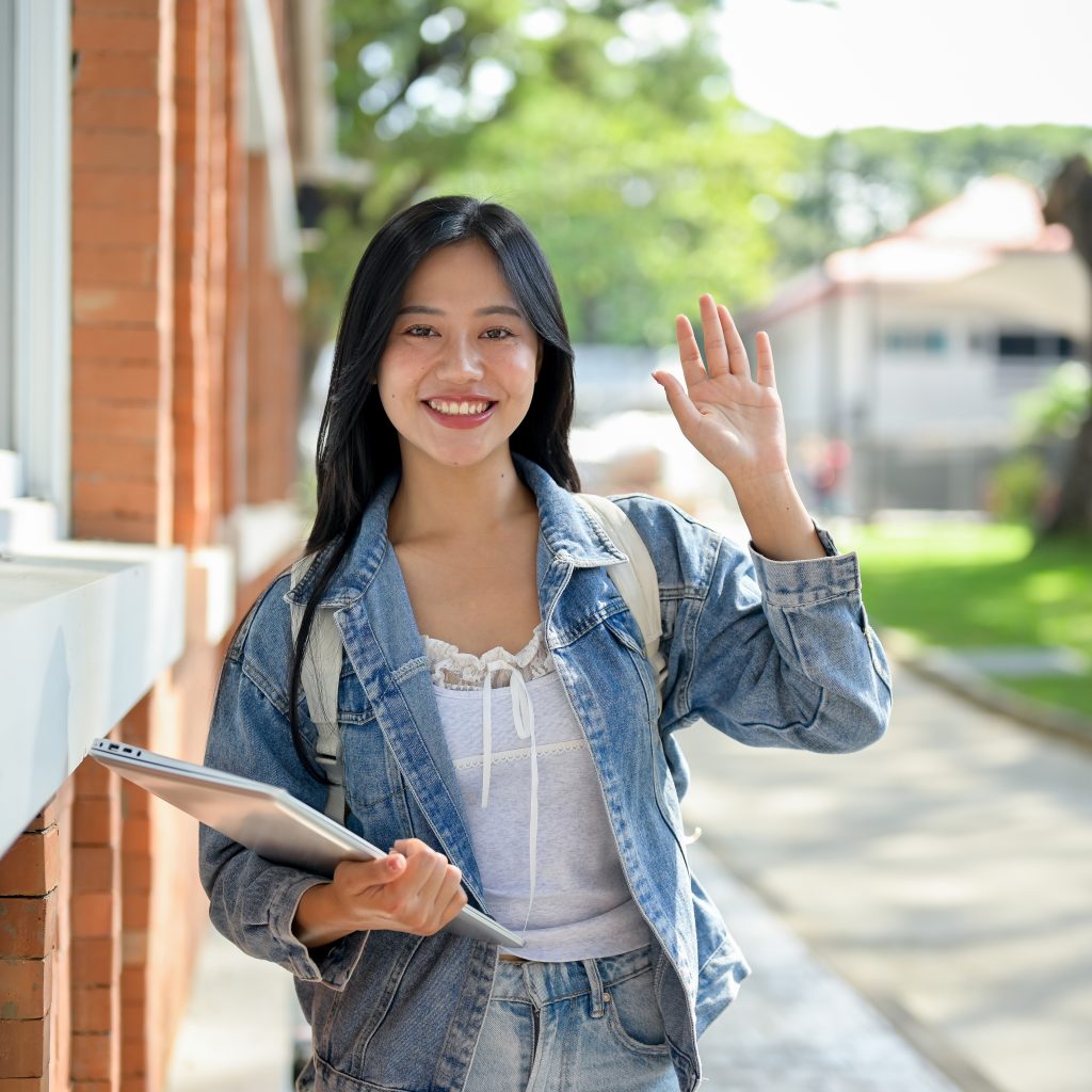 JC Economics student smiling and waving outside a campus building, carrying a laptop and ready to attend her first JC Economics tuition lesson.