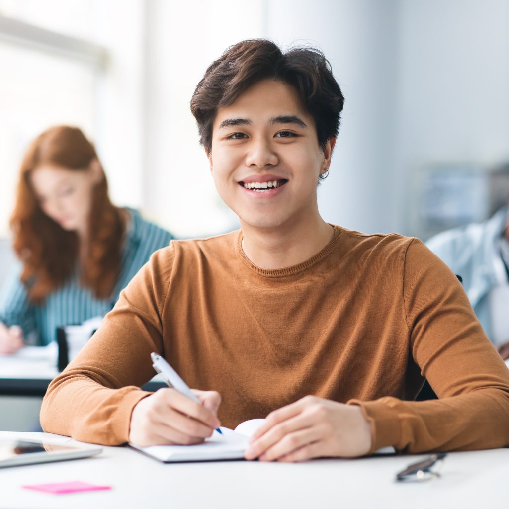 Student smiling while studying in class, focused on JC Economics learning and exam preparation.
